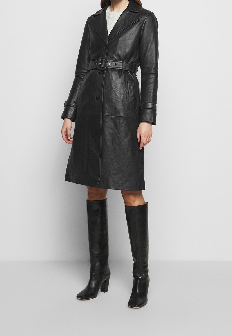 Black leather trench coat with a belted waist, wide lapels, and button closure, paired with knee-high black heeled boots.