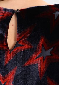 Tejido de terciopelo negro con patrones de estrellas rojas y grises, con una abertura en forma de llave en la parte posterior y un cierre decorativo con botón.
