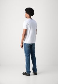 Camiseta blanca de manga corta con cuello redondo, combinada con jeans de mezclilla azul medio y zapatos negros, vista desde atrás.