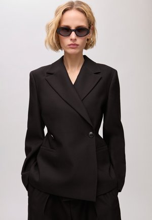 Femme blonde portant un blazer noir sur mesure et un pantalon assorti, des lunettes de soleil noires œil de chat et des boucles d'oreilles créoles dorées, debout les mains dans les poches.
