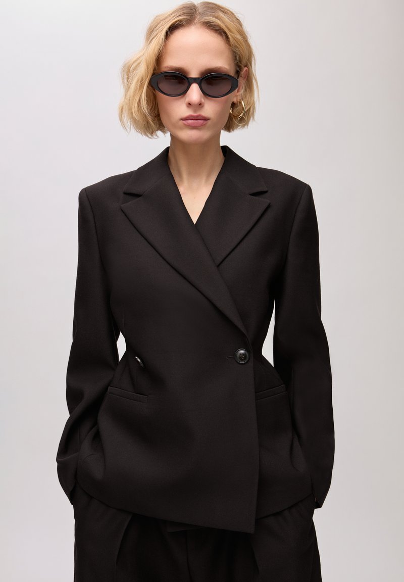 Femme blonde portant un blazer noir sur mesure et un pantalon assorti, des lunettes de soleil noires œil de chat et des boucles d'oreilles créoles dorées, debout les mains dans les poches.