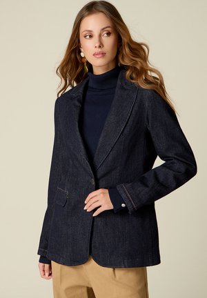 Donna con capelli lunghi e mossi che indossa un blazer di denim scuro sopra un dolcevita blu navy e pantaloni beige, che guarda di lato contro uno sfondo semplice.