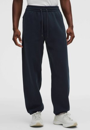 Mand iført navy blå joggingbukser med elastiske manchetter og snøre i taljen, kombineret med hvide sportssko og en hvid skjorte.