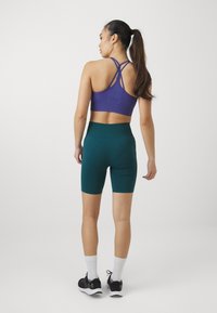 Fioletowy stanik sportowy z krzyżującymi się ramiączkami, połączony z turkusowymi szortami rowerowymi. Ubranie charakteryzuje się dopasowanym krojem oraz gładką, elastyczną teksturą.