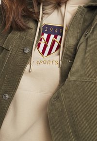 Giacca di velluto a coste verde oliva sovrapposta a una felpa beige. La felpa presenta un logo a scudo rosso e bianco con il testo ricamato "SPORTS."