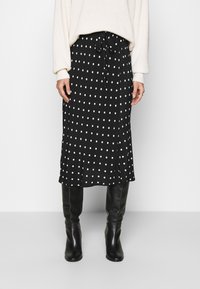 Jupe midi noire à pois blancs, avec taille à cordon de serrage et une légère fente sur le côté, associée à des bottes noires montantes. Tissu doux et coupe décontractée.