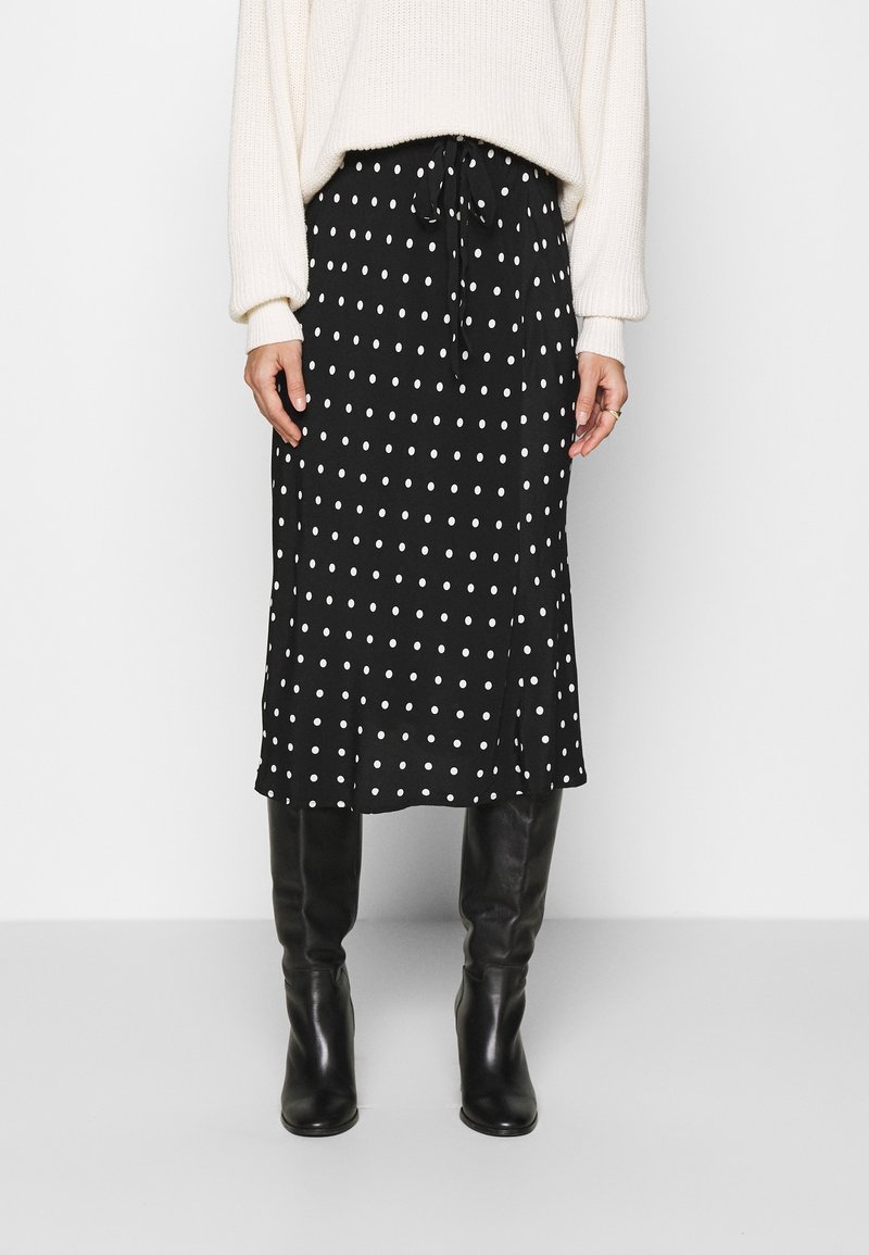 Jupe midi noire à pois blancs, avec taille à cordon de serrage et une légère fente sur le côté, associée à des bottes noires montantes. Tissu doux et coupe décontractée.