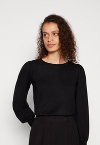Svart langermet genser med rund hals og puffede ermer. Glatt tekstur og tettsittende design, kombinert med høy midje svarte bukser.