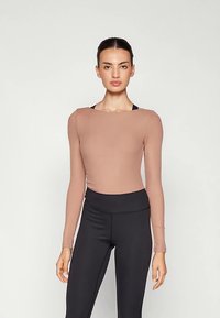 Világosbarna, bordázott hosszú ujjú felső négyzet alakú nyakkivágással. Magasderekú fekete leggingsszel párosítva. Sima textúra, testre szabott fazon.