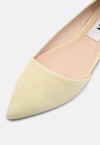 Chaussure plate à bout pointu en daim jaune pâle avec une semelle intérieure beige clair et une étiquette près du talon.