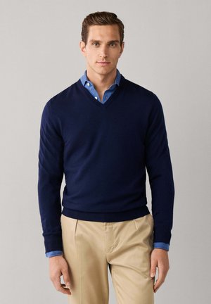 Junger Mann, der einen navyblauen V-Ausschnittpullover über einem blauen Hemd mit Knöpfen und beiger Hose trägt, steht vor einem einfarbigen hellgrauen Hintergrund.