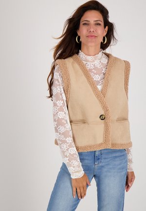 Vrouw in een beige schapenvachtvest over een witte bloemenkant lange mouw top en blauwe spijkerbroek, met gouden hoepel oorbellen.