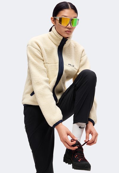 Νέος άνθρωπος με μπεζ fleece μπουφάν και μαύρο παντελόνι δένει μαύρο μπότες πεζοπορίας ενώ φορά ανακλαστικά μεγάλα γυαλιά ηλίου.