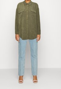 Camisa de pana verde oliva con cuello, cierre frontal de botones y dos bolsillos en el pecho. Combinada con jeans rectos de mezclilla azul claro y sandalias blancas.
