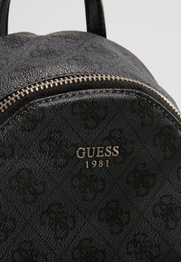 Sac à dos noir texturé Guess avec fermeture éclair argentée et logo en métal indiquant "GUESS 1981" sur la poche avant.