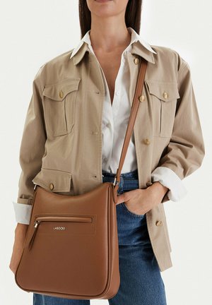 Kobieta w beżowej kurtce i białej koszuli z niebieskimi dżinsami, niosąca brązową skórzaną torbę na ramię z przednią kieszenią na zamek.