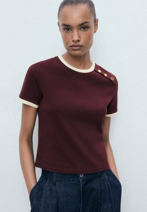 Burgundy short-sleeve top with a cream-colored ribbed neckline and sleeve edges; features gold buttons on the left shoulder.