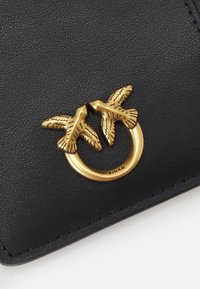 Svart teksturert skinnlommebok med en gullfarget metalllogo som har to fugler som danner en hjerteform og en sirkulær ring.