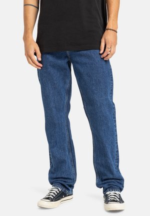 Jean en denim bleu moyen avec une coupe décontractée, comportant cinq poches et un léger dégradé. Associé à des baskets noires montantes.