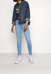 Chaqueta de mezclilla en azul oscuro con las mangas arremangadas, camiseta negra con un diseño multicolor, jeans ajustados azul claro y zapatillas altas blancas.