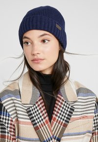 Knit navy beanie featuring a ribbed texture and a small branded tag. Worn with a multicolored plaid coat in beige, black, and red tones.