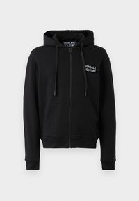 Must mustaga kapuutsiga must sweatshirt, millel on ees tasku ja valge Versace Jeans Couture logo vasakul rinnal. Pehme kangastekstuuri.