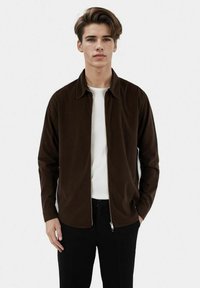 Brown corduroy jacket with a zip front, collar, and long sleeves, worn over a white T-shirt, paired with black trousers.