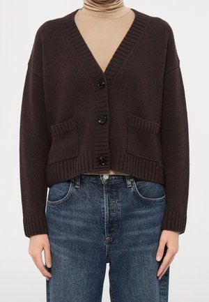 Mørkebrun strikket cardigan med tre sorte knapper, frontlommer, båret over en beige rullekrave og kombineret med blå jeans.