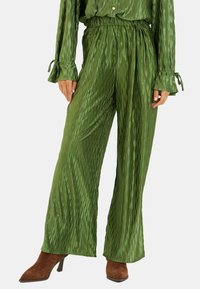 Pantalones anchos plisados verdes hechos de un tejido brillante con una cinturilla elástica y líneas verticales texturizadas. Combinados con botas de tacón marrones.