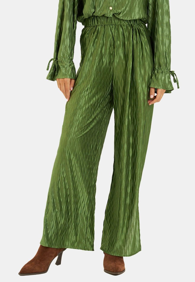 Pantalones anchos plisados verdes hechos de un tejido brillante con una cinturilla elástica y líneas verticales texturizadas. Combinados con botas de tacón marrones.