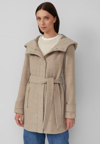 Manteau en laine beige avec une grande capuche, une ceinture et des poches latérales zippées. Tissu texturé, coupe droite et manches longues avec détails en bouton.
