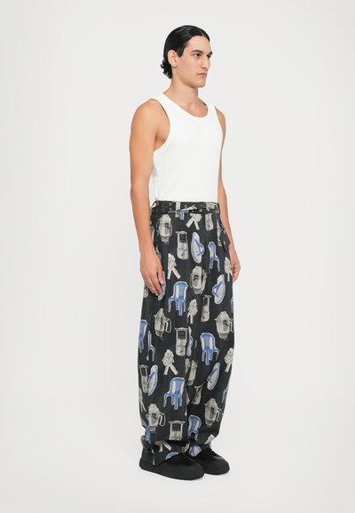 Pantalons noirs avec un motif unique de chaises et d'objets bleus, à coupe loose et avec une ceinture contrastante. Portés avec un débardeur blanc.