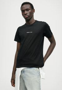 Camiseta de algodão preta com um logótipo branco "REPLAY" no peito. Manga curta e Design de gola redonda. Usada com jeans azuis desgastados.