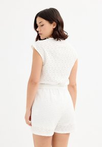 Mono blanco con detalles de ojal, que presenta una cintura ajustada, mangas cortas y un patrón texturizado con recortes, ofreciendo un diseño ligero y transpirable.
