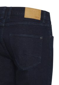 Sötét denim farmer, sima textúrával, öt zsebes dizájnnal és barna bőr folttal, amelyen az "ISOLID" márkázás található a deréknál.