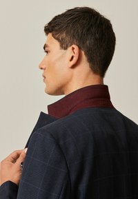 Veste ajustée en tissu noir marin foncé avec un motif à carreaux. Comprend un col bordeaux contrastant, une finition texturée et des détails subtils.
