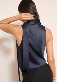 Tengerészkék szatén ujjatlan felső, magas gallérral és hátul egy hosszú szalaggal megkötve, sima textúrával és testhezálló szabással.