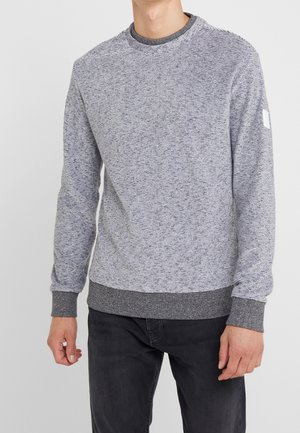 Mann trägt einen grau-weiß gesprenkelten Langarmpullover mit dunkelgrauen Bündchen und Saum, kombiniert mit schwarzer Hose, stehend.