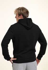 Sudadera negra de algodón con una textura suave, cuenta con una capucha con cordón y puños acanalados, mostrada desde atrás con pantalones grises.