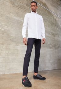 Camicia bianca con bottoni, pantaloni slim-fit scuri e scarpe sportive nere con dettagli rossi e bianchi su un pavimento texturizzato contro un muro di mattoni.