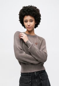 Pruun soonikkoega kampsun tekstuurse viimistlusega ja ümara kaelusega, vasakul küljel väike ümmargune logo. Kandmisega mustad püksid.