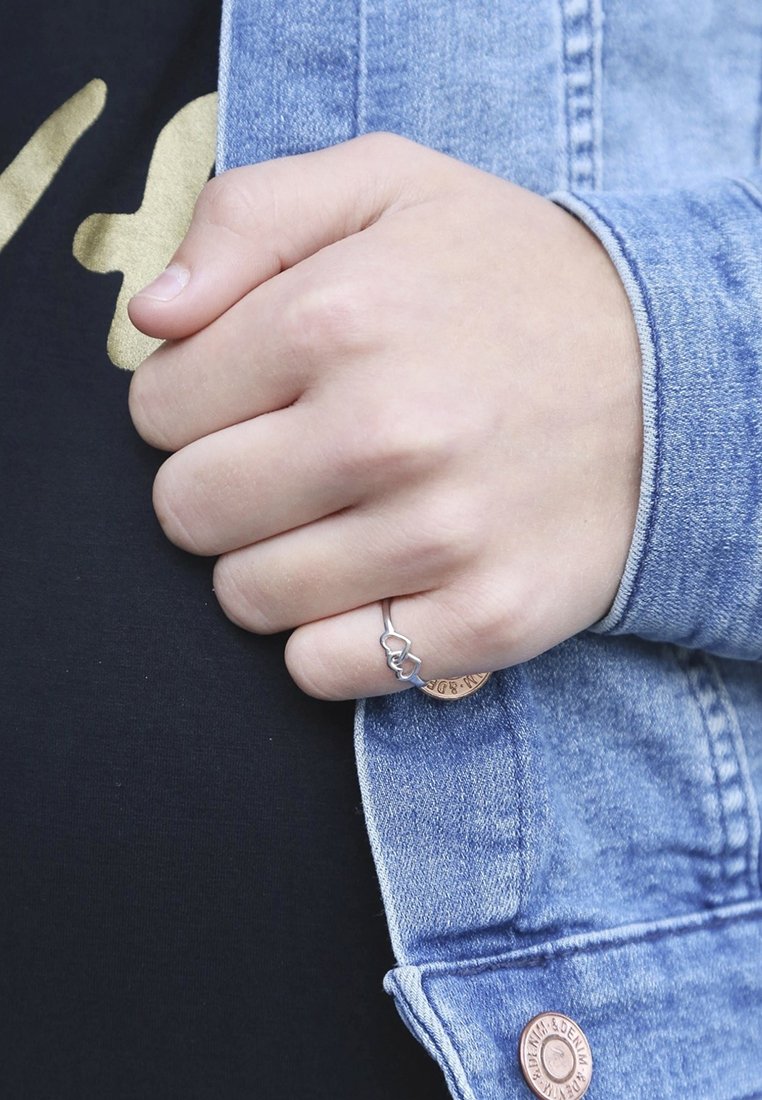 Zilveren ring met een verstrengeld hartontwerp op een hand, met een spijkerjack over een zwart shirt. Achtergronddetails zijn minimalistisch.