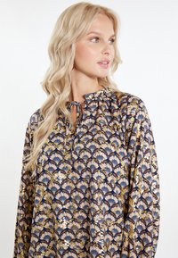 Blusa a maniche lunghe in navy con un motivo floreale caratterizzato da accenti oro, blu e rosa. Collo arricciato con dettaglio a lacci. Tessuto morbido.