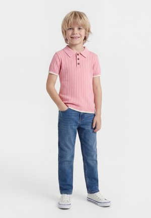 Ragazzo giovane sorridente con capelli biondi che indossa una polo rosa a coste, jeans blu e scarpe da ginnastica bianche, in piedi con una mano in tasca.