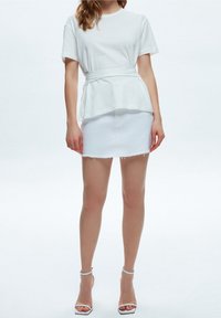 White cotton t-shirt with peplum waist, paired with a white denim mini skirt featuring frayed hem. Model wears clear heeled sandals.