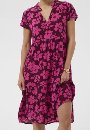 Femme portant une robe violette avec de grands motifs floraux rose vif, manches courtes et un col volanté, tenant l'ourlet d'un côté.