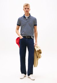 Marinrandig polo skjorta, marinblå byxor, vita sneakers. Röd keps i handen, beige jacka draperad över armen. Enkel och avspänd design.
