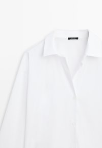 Chemise blanche à manches longues avec un col pointu et des boutons sur le devant, comportant une étiquette noire cousue à l'intérieur de l'encolure.