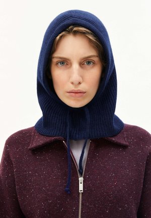Frau mit blauer Strickhaube, die eng um den Kopf gezogen ist, trägt einen weinroten Reißverschlussmantel mit Kragen.