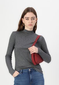 Hanorac gri cu mâneci lungi și guler înalt, accesorizat cu o geantă roșie pe umăr. Textură fină, design ajustat, pe un fundal alb.